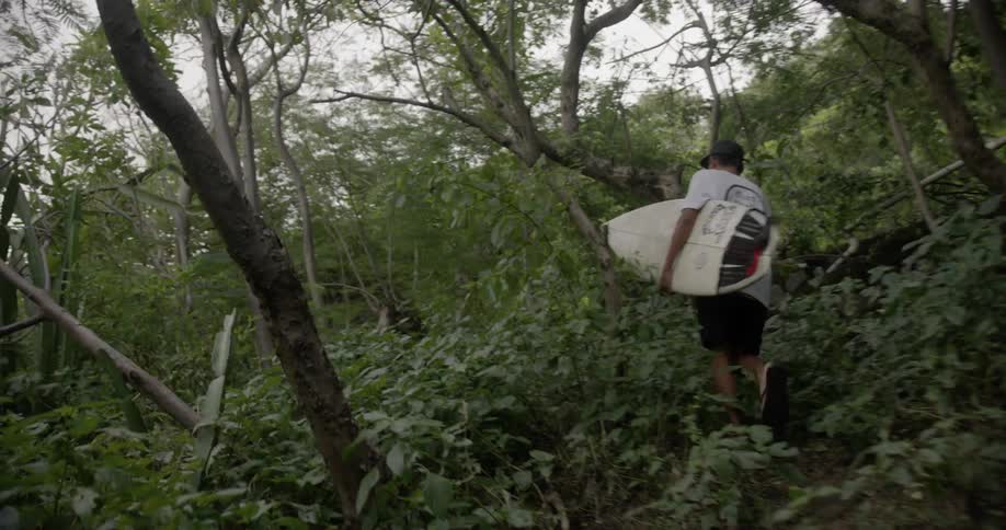 Surfer carrying his surfboard hiking up a trail through the jungle