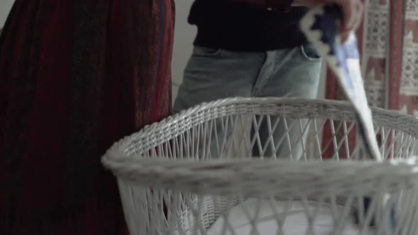 A male holding a white stuffed rabbit closely to his chest watches a pregnant female lay a crocheted blue and white blanket into a white wicker bassinet