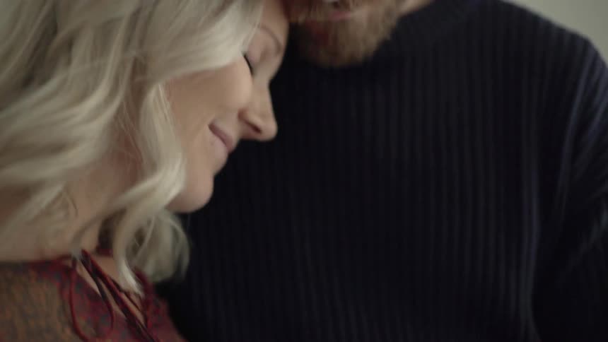 A bearded man kisses his blonde haired pregnant wife as they embrace and each place a hand to feel the movements of their unborn child