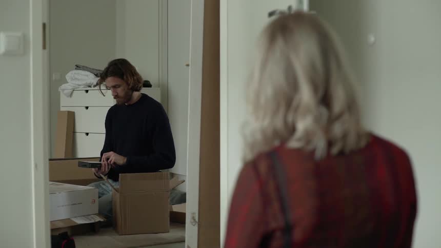 A woman stands looking through a doorway where a man sits on the floor surrounded by boxes and holds an item up to her