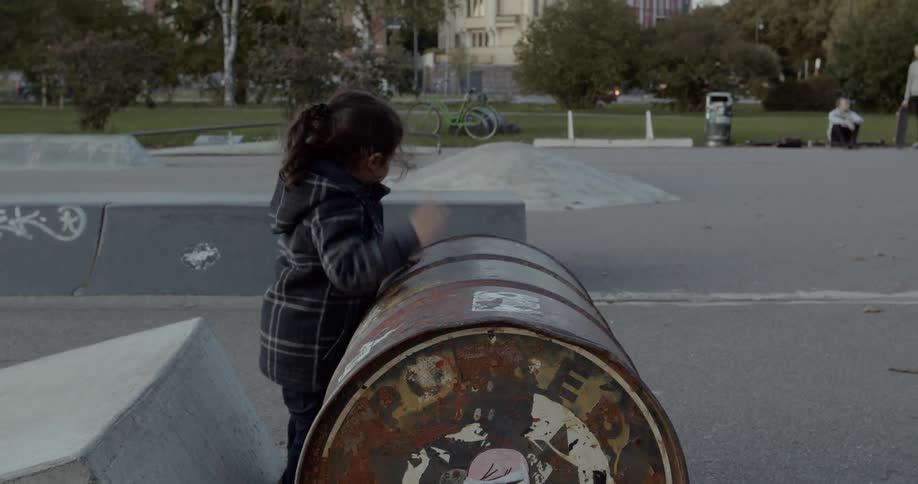A young girl in a skate park playing drums on a sideways metal barrel