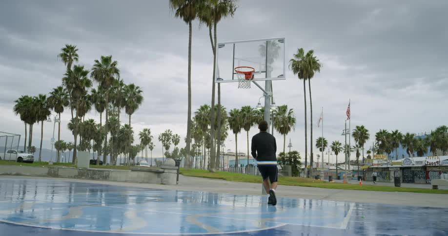A man dribbles a basketball between his legs a few times before performing a layup shot and sinking the ball in the hoop on an outdoor basketball court