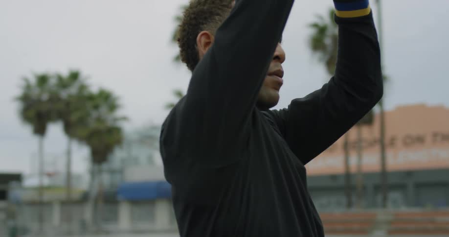 A man stops dribbling a basketball to take a shot on a basketball court with buildings and palm trees in the back ground