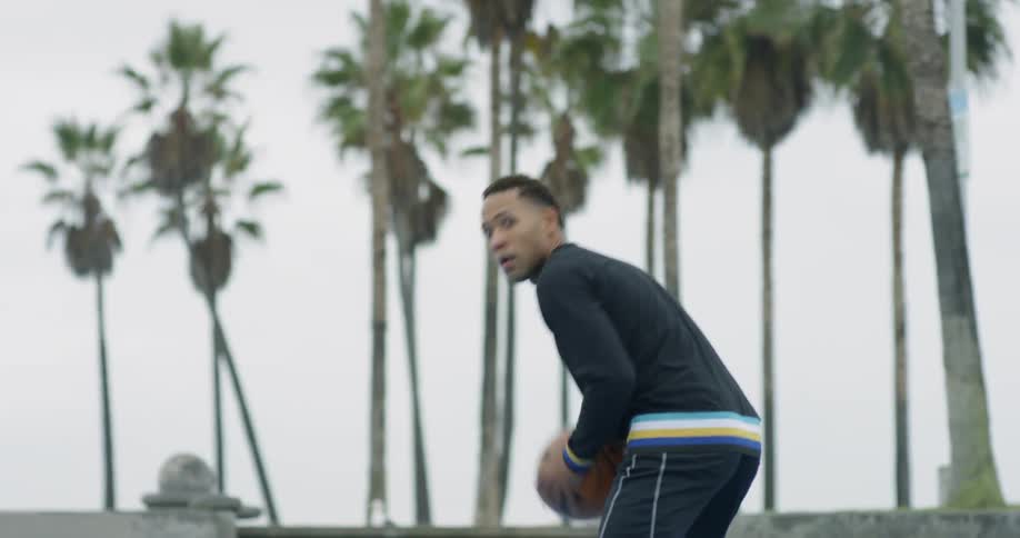 A black male dribbles a basketball on an outdoor basketball court before shooting it at the hoop