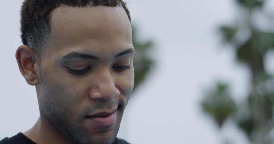 A young black man stands outdoors and smiles to himself