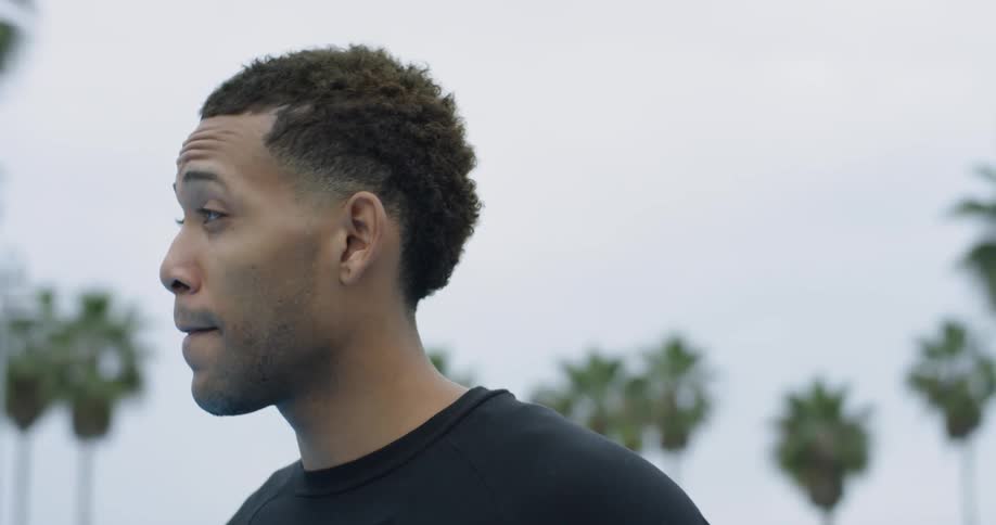 A man holding a basketball takes a deep breath while looking around while seagulls fly behind him near palm trees