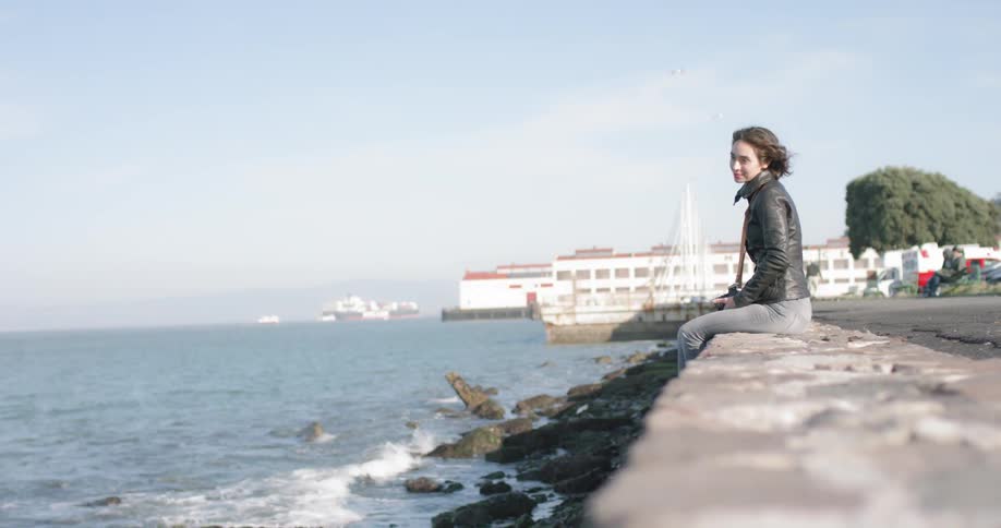 A female sits on the edge of a strip of pavement looking out over the ocean and then snaps a picture