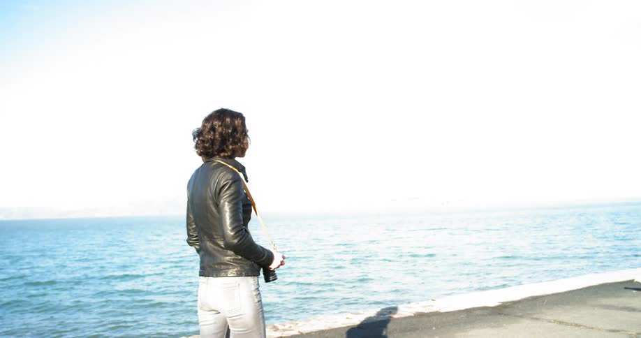 A female walks along the edge of a body of water and raises her camera to snap a picture of a bird in front of her
