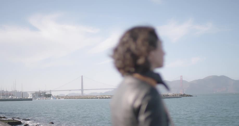 Woman stands near ocean with suspension bridge and mountains on horizon