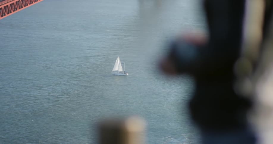A female with a camera on a strap around her neck stands at the edge of a pier watching a sailboat in the ocean below with a bridge across from her