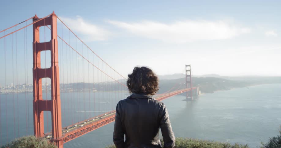 A female wearing a dark jacket stands from an elevated position looking out over a bridge and body of water
