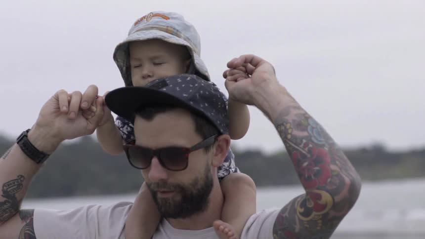 A man has a baby on his shoulders as he stands by the water on a cloudy day