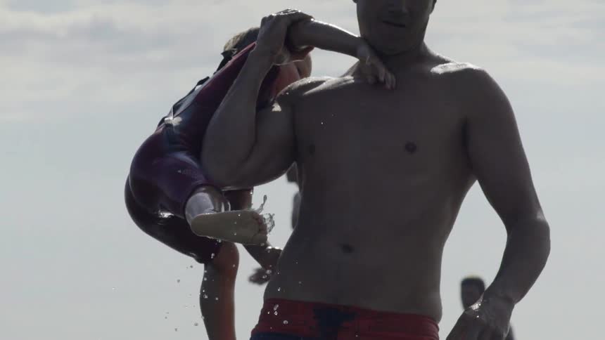 A man lifts a boy in a wetsuit out of the water and onto his back