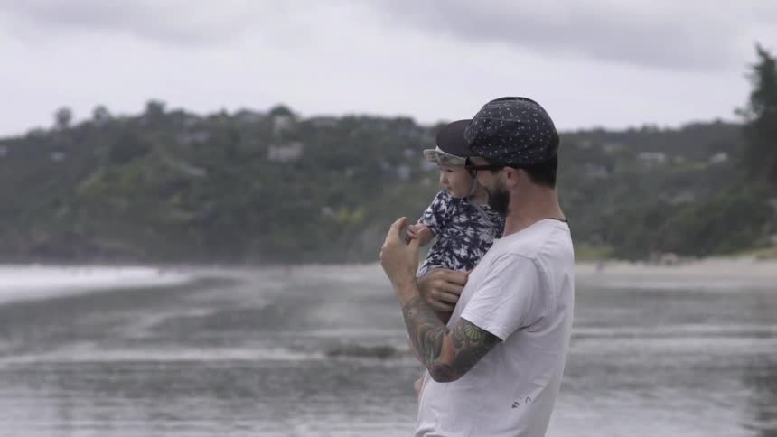 A man is holding a baby by the water on a cloudy day