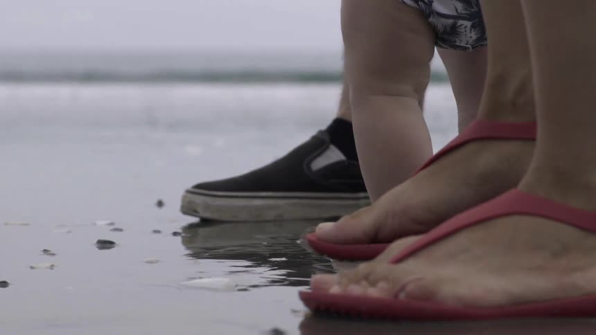 A baby's bare feet, and two sets of adult feet, take steps on a wet beach