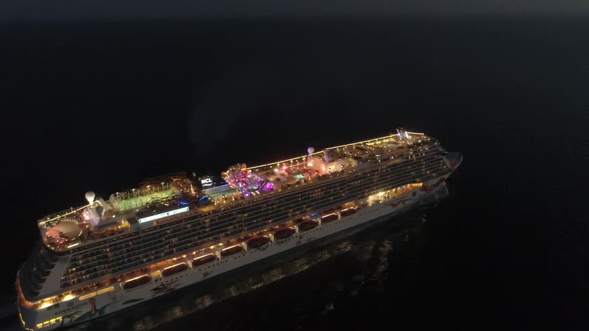 A large cruise ship with a pool on the top deck is brightly lit as it sails through the ocean at night