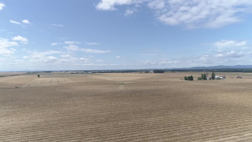 The plowed brown fields of a farm lie under a blue sky with scattered clouds