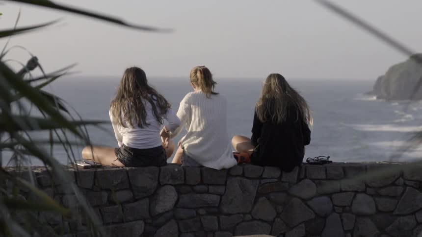 Three women sit on a stone wall overlooking the ocean