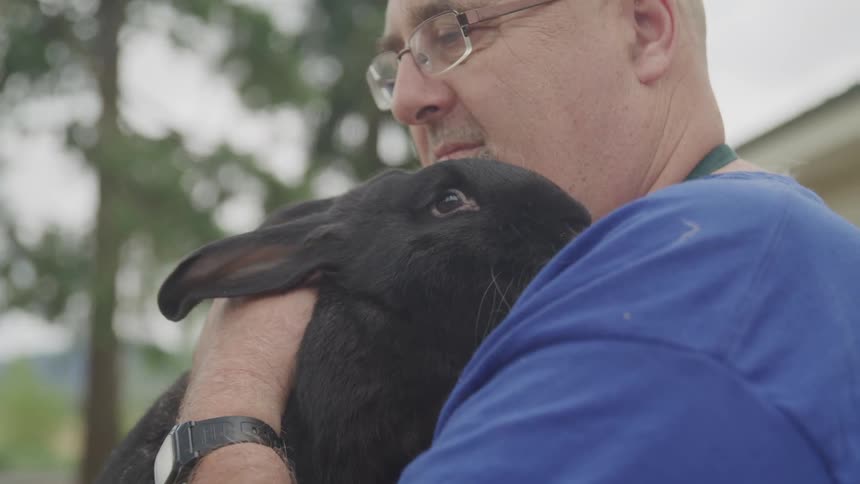A white man wearing glasses is holding a large black rabbit and petting it behind the ears