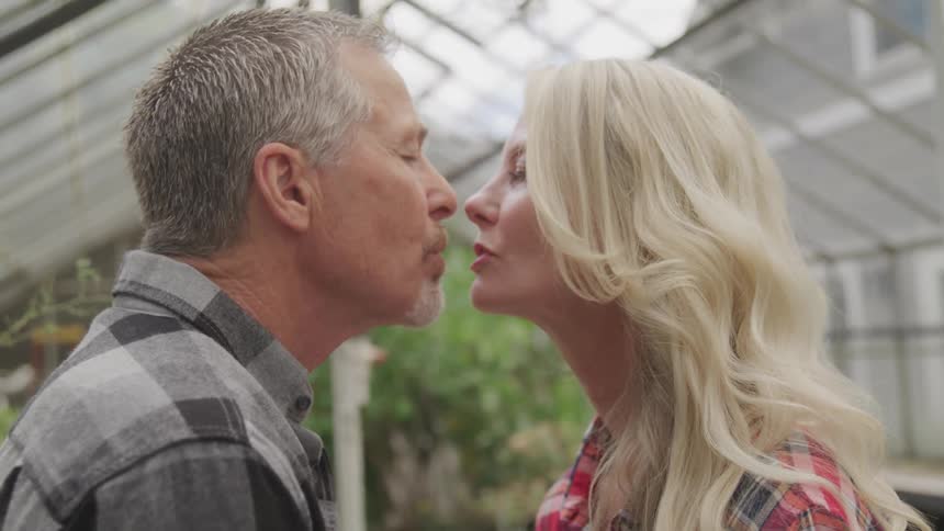 A middle aged white man and white woman go in for a kiss in a greenhouse