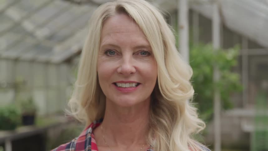 A blonde woman stands in a greenhouse and smiles