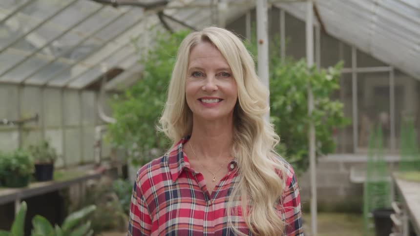 A woman with long blonde hair smiles while standing in a greenhouse