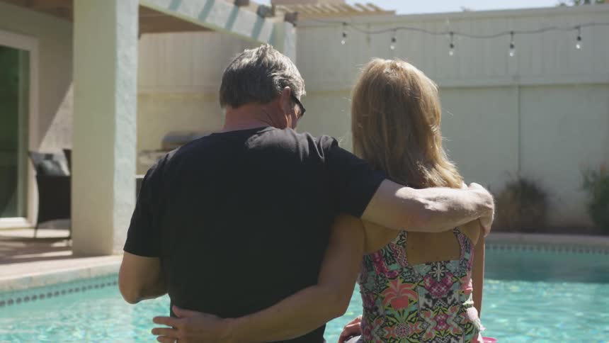 A man and woman hug while sitting next to an outdoor swimming pool