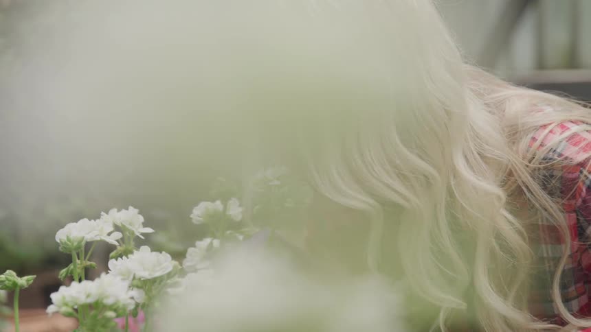 Blonde woman wearing gardening gloves smells a flower in her garden