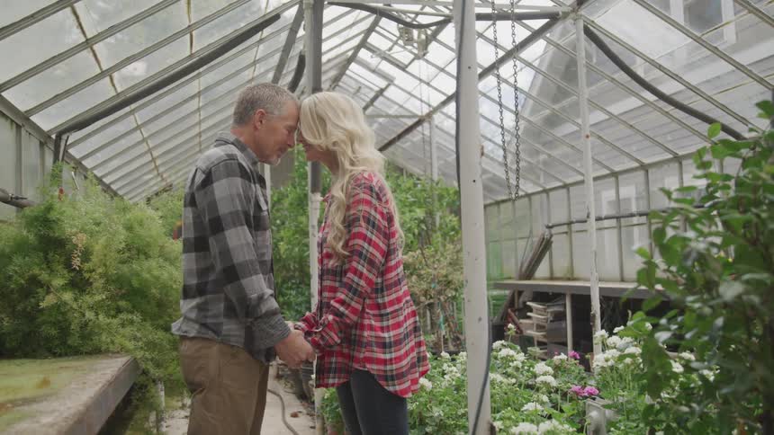 A man and a woman stand in a greenhouse holding hands