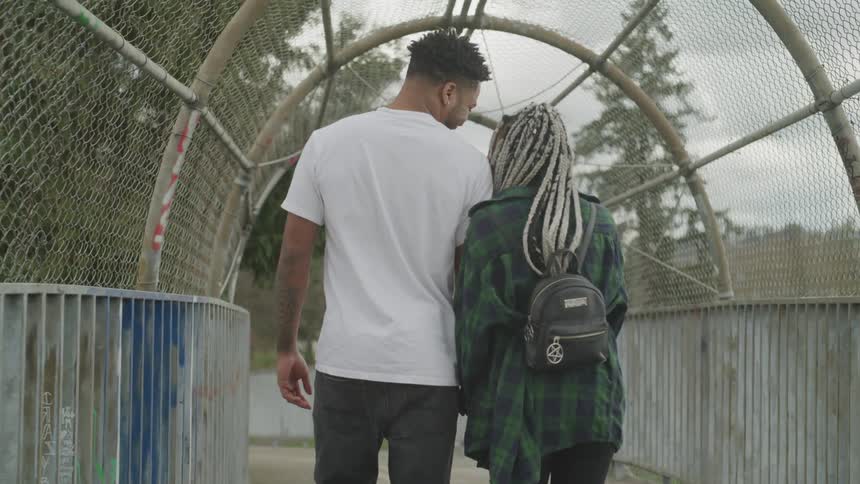 A man and woman snuggle closely walking on a covered pedestrian bridge