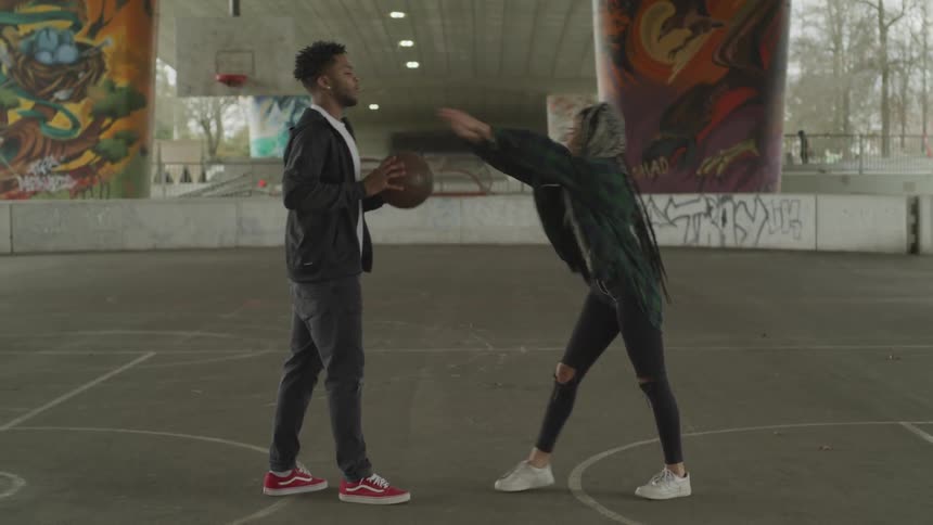 A young black man and woman playfully fighting over a basketball on a concrete basketball court under a bridge