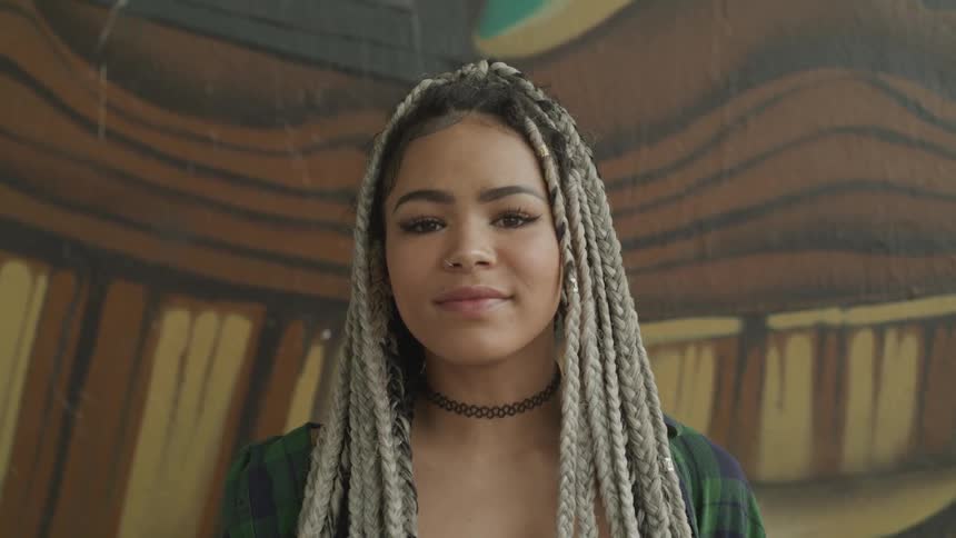 A woman with braided hair stands posing in front of a mural on a wall