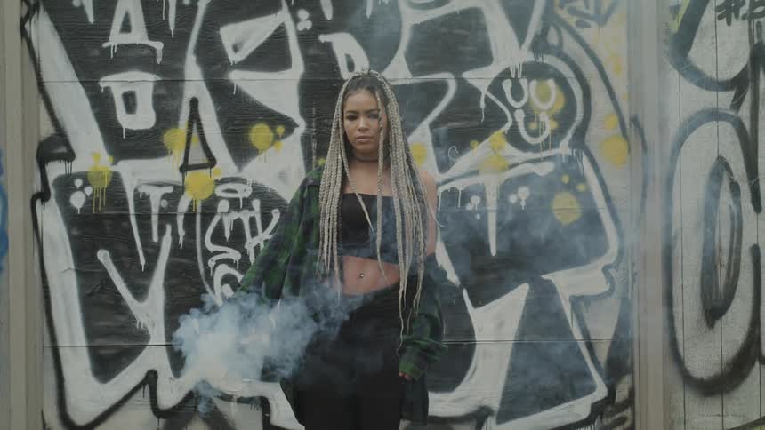 A woman holding a smoke stick stands in front of a graffiti covered wall