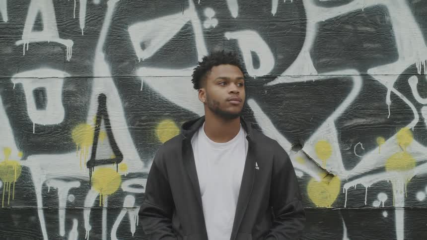 A man is standing next to a wall with his hands in his pockets looking left to right