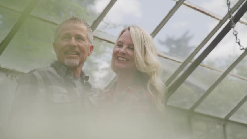 A man and woman in plaid shirts smile while standing in a greenhouse
