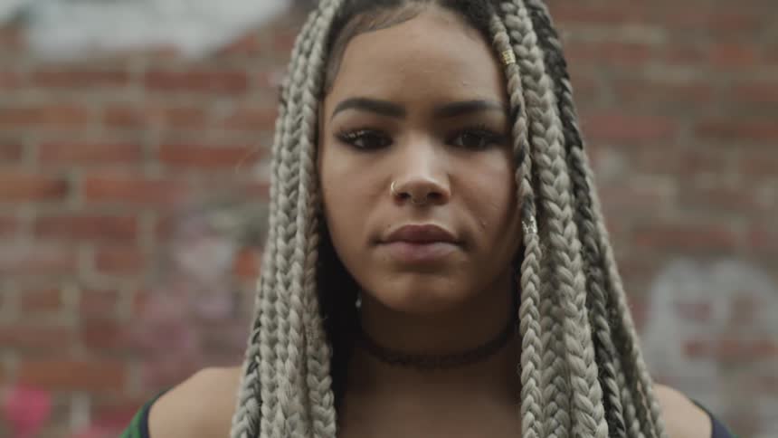 Teenage girl with long braids and a choker necklace lifts her head and looks into the camera as she stands outside in front of a brick wall