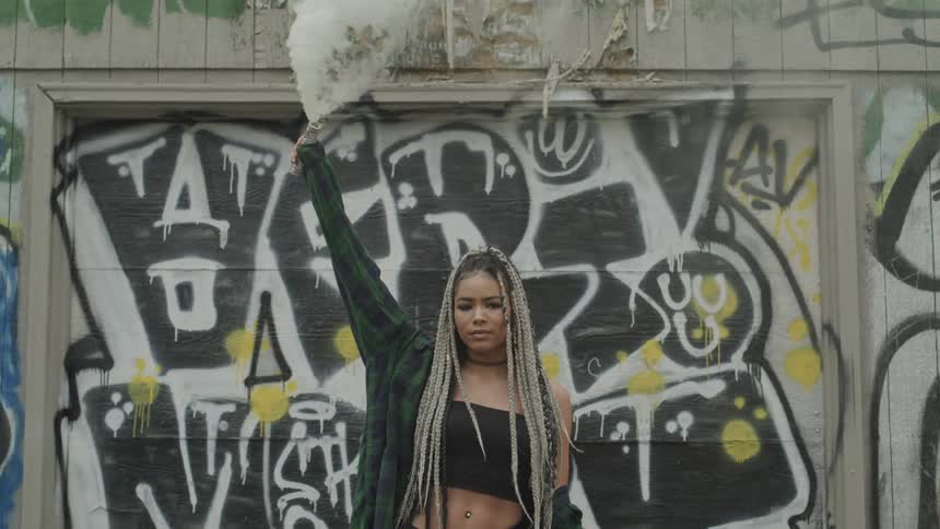 A young woman is holding up a smoking canister while standing in front of graffiti