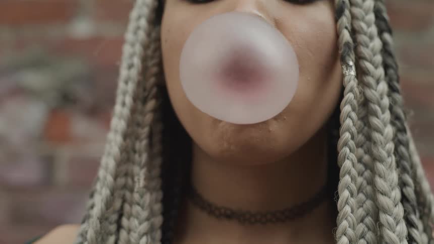A woman with long braids blows a bubble until it pops on her lips
