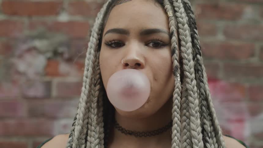 A woman blows a bubble in front of a brick wall