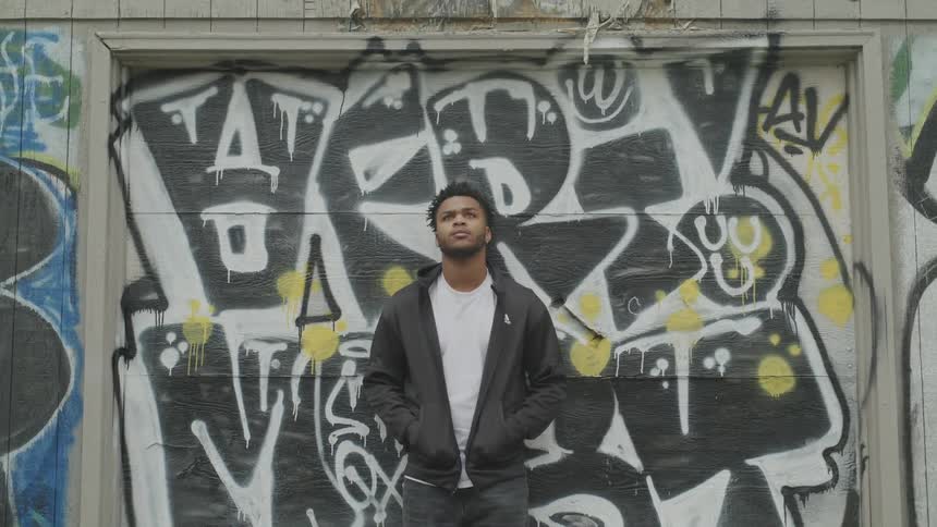 A young man stands in front of a grafitti filled wall