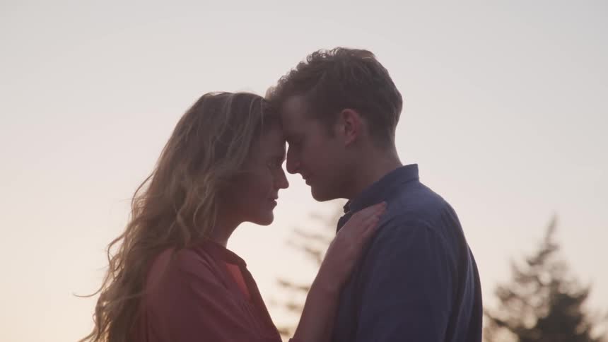 A man and woman stand together with their foreheads touching in a loving embrace in front of some trees and the setting sun
