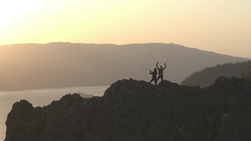 The silhouette of two hikers holding up their poles in triumph as they stand on a rocky ridge at sunset