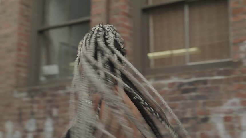 A female in front of a brick building with graffiti twirls around once to the right and back to the left as her long braids swing around with her