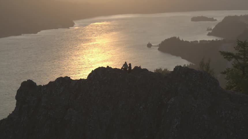 Couple sitting on top of mountain peak overlooking a lake at sunrise