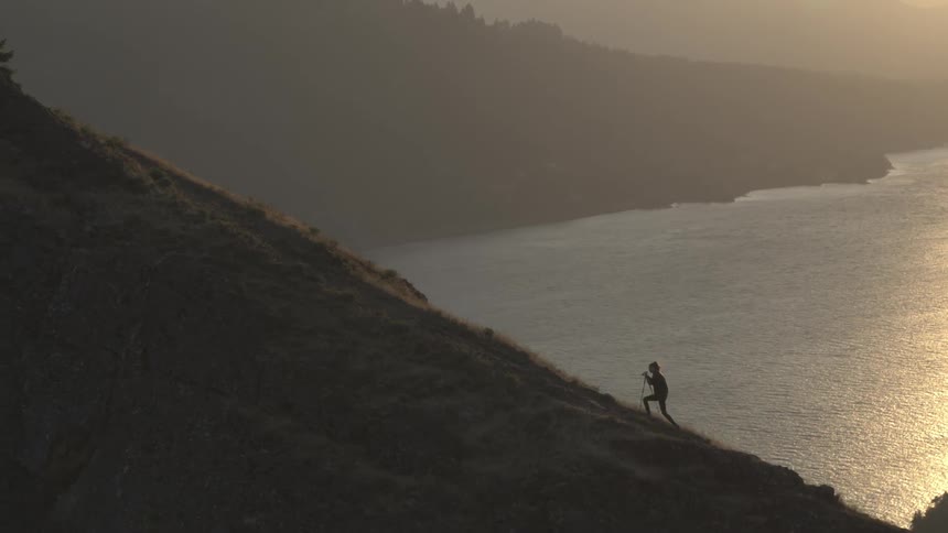 A woman briskly hikes up a mountain that is located along a mountainous coastline and the ocean is seen far below