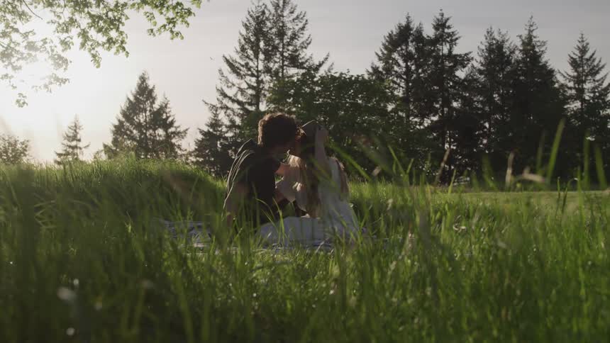Man and woman sit on the grass and kiss each other by late afternoon on a sunny day
