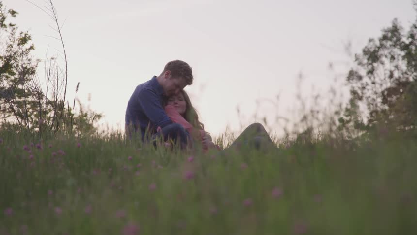 A man sitting in a field embracing a woman who is laying against his chest