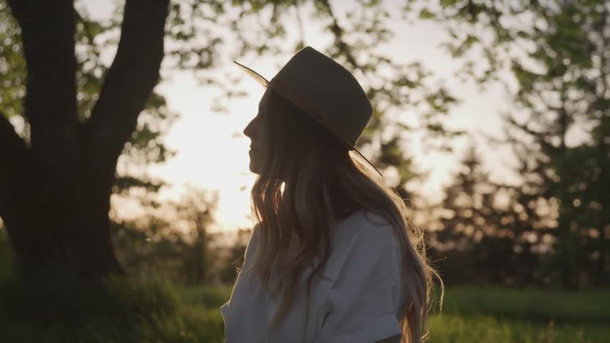 Blonde woman wearing wide hat looks ahead in the woods at sunset