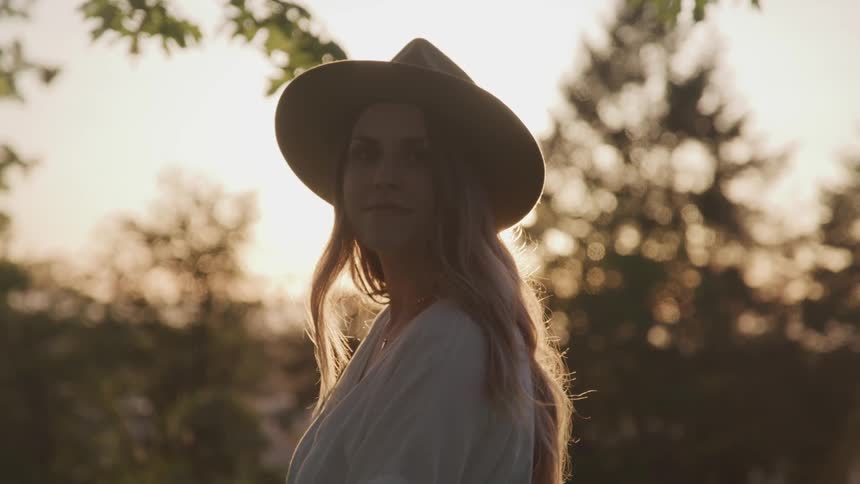 Woman wearing wide hat looks at the camera and smilesin the forest while the sun goes down