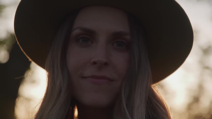 A woman in a hat looks into the camera at sunset